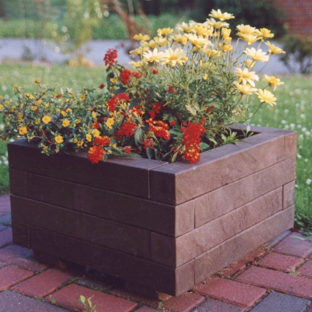 brauner quadratischer Blumenkübel mit Blumen bepflanzt auf Gehweg vor Wiese