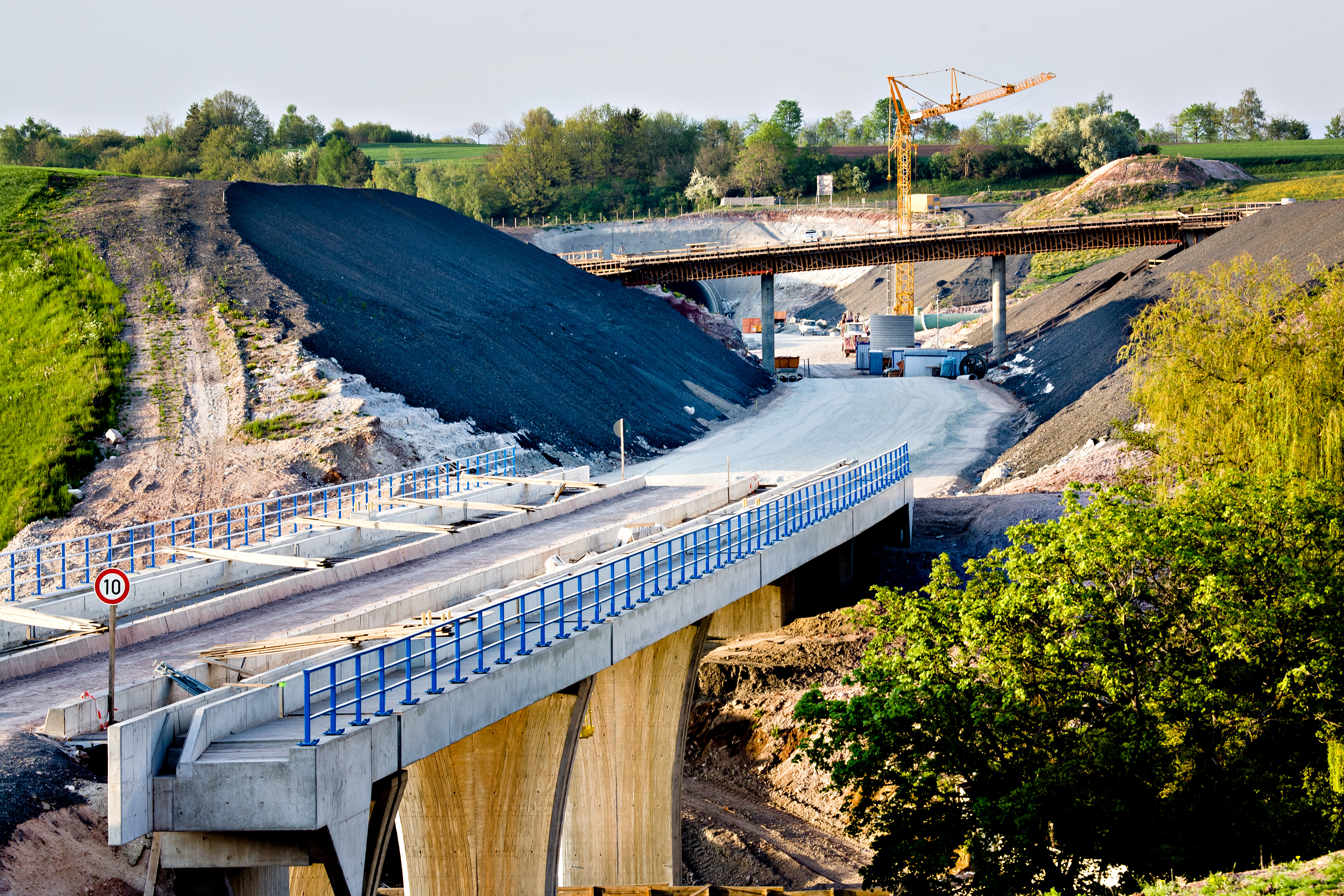 Bau einer Brücke mit einer Baustelle im Hintergrund. Sichtbare Materialien sind Asphalt und Geotextilien. Ein Kran ist im Einsatz.