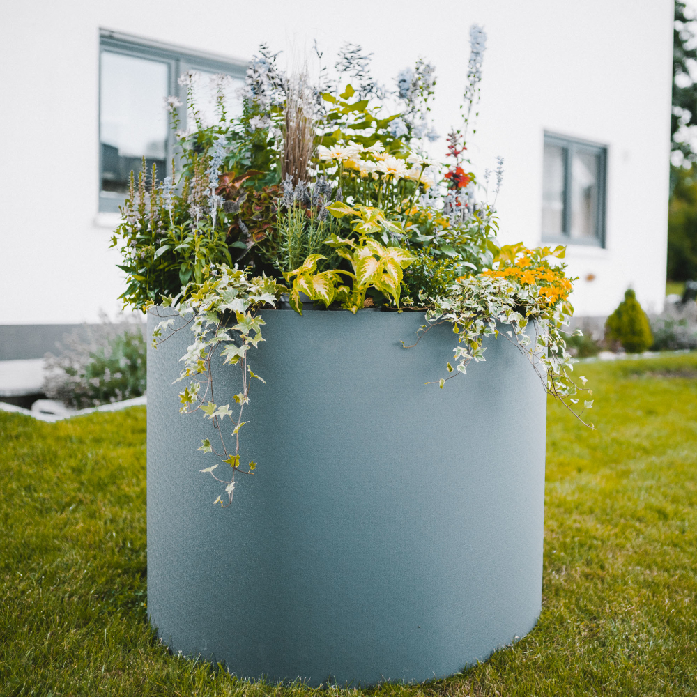 rundes hellgraues Hochbeet mit Pflanzen auf Wiese vor Haus