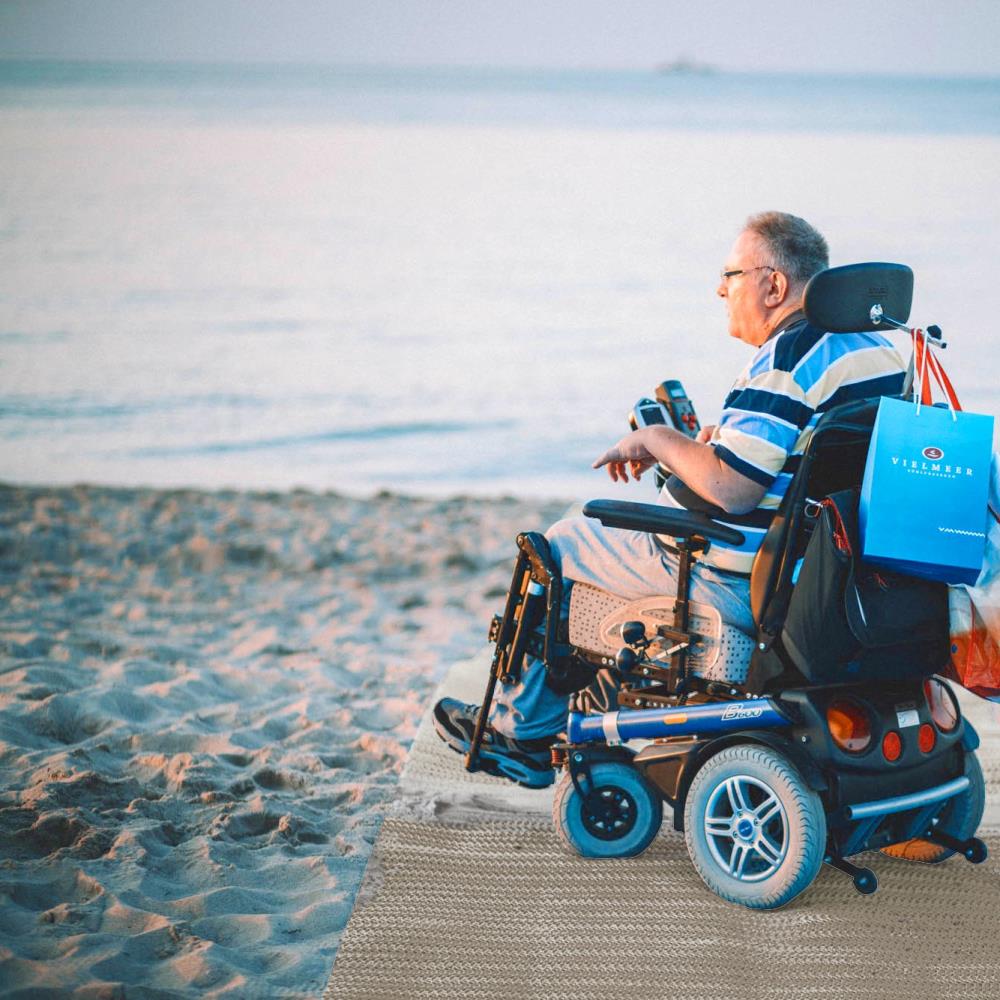 Rollstuhlfahrer auf Sandmatte mit Meer im Hintergund.