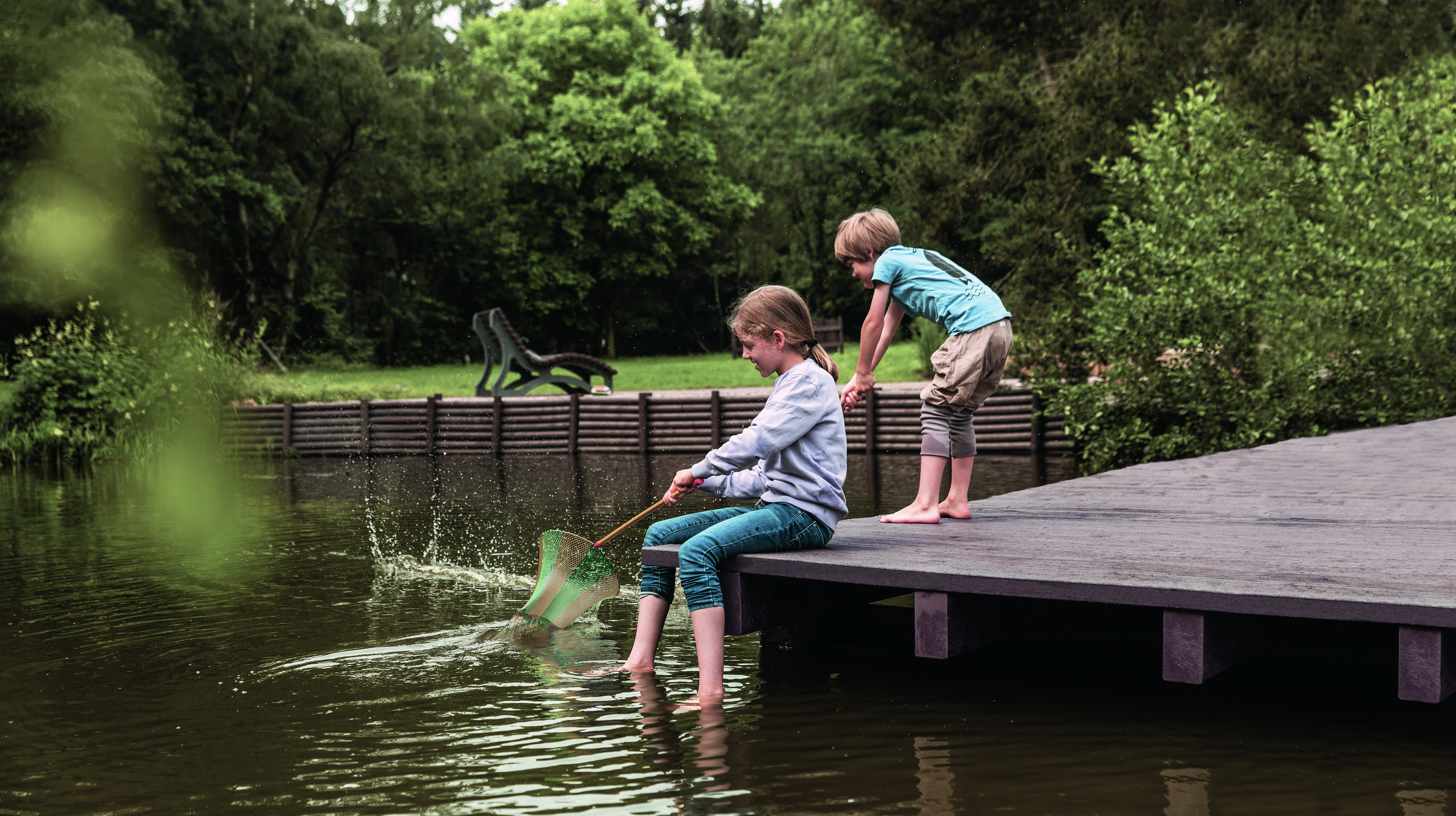 hanit Recyclingkunststoff Zwei Kinder stehen auf einem Steg aus hanit Recyclingkunststoff und fangen mit einem Netz Fische im Wasser. Im Hintergrund sind Bäume und eine hanit Bank sichtbar.