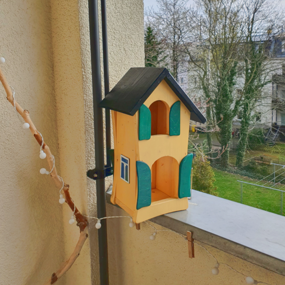 gelbes Vogelhäusschen an einem Balkon montiert