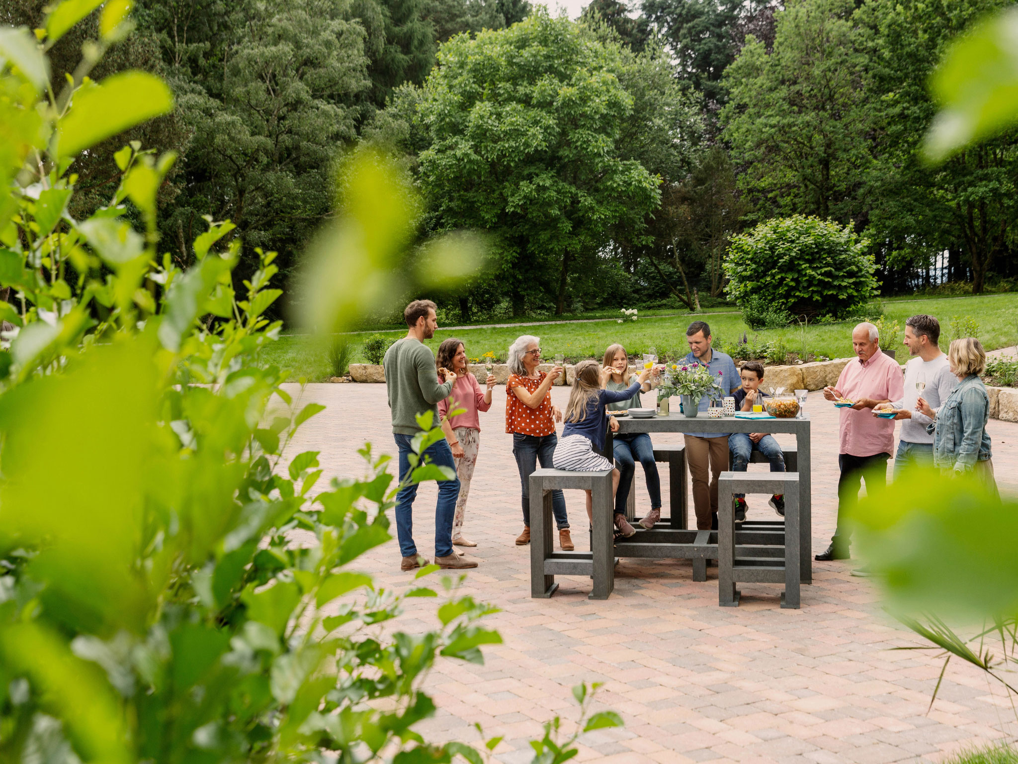 Gruppe von acht Personen, die um einen Tisch mit hanit Gartenmöbeln aus Recyclingkunststoff stehen. Im Hintergrund Bäume und Rasen.